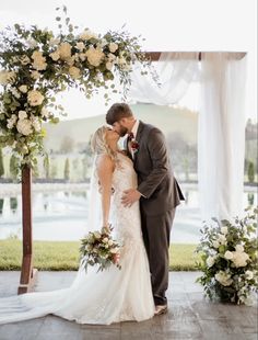 a bride and groom kissing under an arch decorated with white flowers at their wedding ceremony