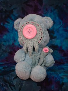 a gray stuffed animal with a pink button on it's head sitting in front of a blue and purple background