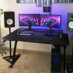 a computer desk with two monitors and speakers on top of it in front of a purple wall