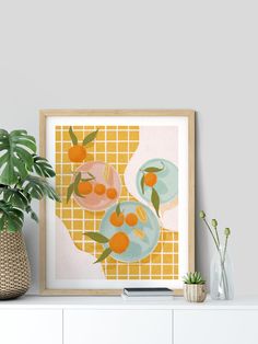 an art print with oranges on a yellow and white checkerboard background in a wooden frame next to a potted plant