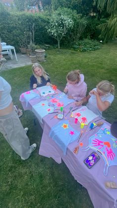 a group of children sitting at a table with paintings on it in the grass outside