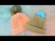 two crocheted beanies with pom - poms sitting on a wooden surface