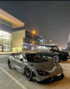 two sports cars parked in front of a building at night with the lights on and buildings lit up behind them