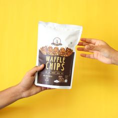 two hands reaching for a bag of waffle chips on a yellow background with one hand holding the packet