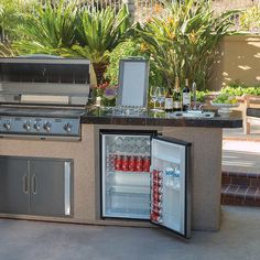 an outdoor kitchen with two refrigerators and a grill in the back ground, surrounded by plants