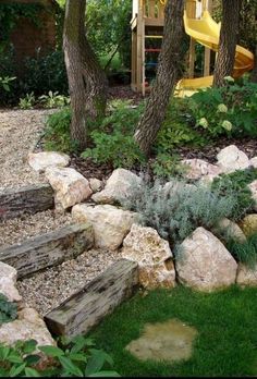 an outdoor play area with rocks, plants and a yellow slide in the background is surrounded by trees