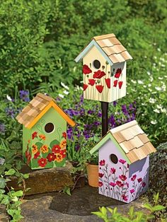 three bird houses are sitting on the ground in front of some flowers and bushes,