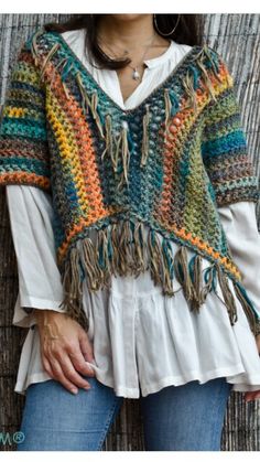 a woman standing next to a wall wearing a colorful crochet shawl with fringes