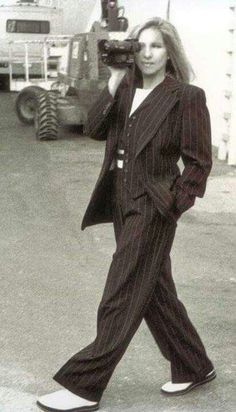 a woman walking across a parking lot while holding a camera to her face and wearing a suit