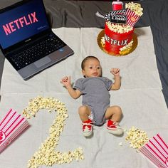 a baby sitting in front of a laptop computer next to popcorn buckets and a cake