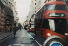 a double decker bus driving down a street next to tall buildings and people walking on the sidewalk