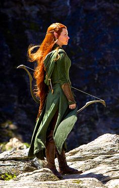 a woman with long red hair is dressed in green and holding a bow on her shoulder