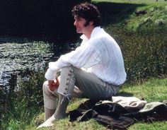 a man in white shirt and pants sitting on grass next to water with his hand up