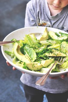 a young boy holding a white bowl filled with lettuce and avocado