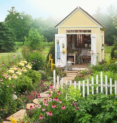 a garden with flowers and a shed in the back ground, next to a white picket fence