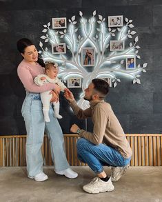 a man and woman holding a baby in front of a tree with pictures on it