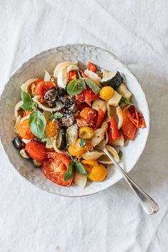 a white bowl filled with pasta, tomatoes and olives on top of a table