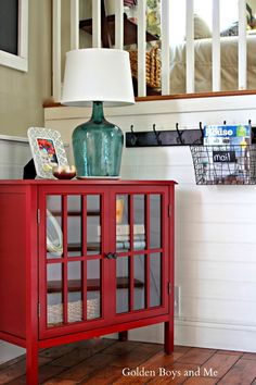 a red cabinet sitting on top of a wooden floor next to a lamp and pictures