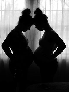 two pregnant women standing next to each other in front of a window with sheer curtains