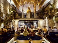 people sitting at tables in a restaurant with stone walls and exposed ceilings, while one person stands on the second floor