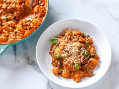 a white bowl filled with pasta next to a blue casserole dish on a marble counter