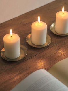 three lit candles sitting on top of a wooden table next to an open book,