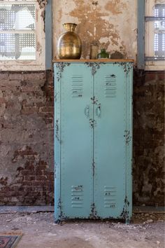 an old blue metal locker in front of a brick wall with windows and a vase on top