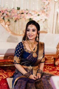 a woman in a blue and gold sari sitting on a red carpet with flowers behind her