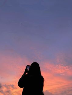 a woman is taking a photo with her cell phone at sunset or dawn, while the moon is in the distance