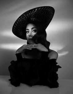 a woman in a black dress and hat posing for the camera with her hands on her chest