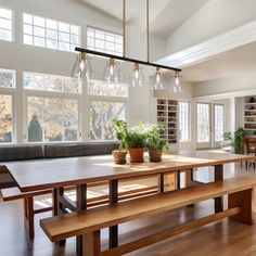 a dining room table with benches and potted plants in the center is surrounded by large windows