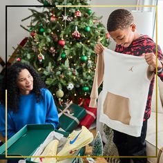 a young boy holding up a white shirt next to a woman in front of a christmas tree