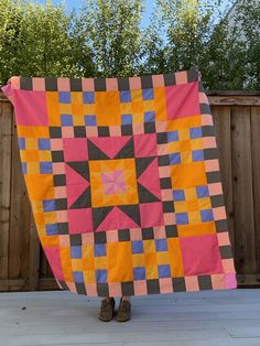 a person standing in front of a fence holding up a large pink and yellow quilt