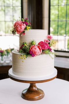 a white wedding cake with pink flowers on top and greenery in the middle, sitting on a wooden stand