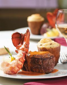 a white plate topped with meat and shrimp next to a glass of wine on top of a table