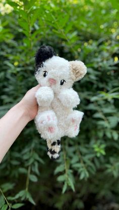 a hand holding a small white stuffed animal in front of some bushes and trees with green leaves