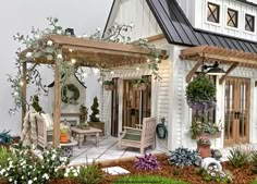 a small white house with lots of plants and flowers around the front porch, covered by a pergolated roof