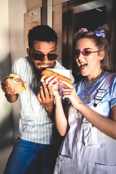 a man and woman standing next to each other holding sandwiches