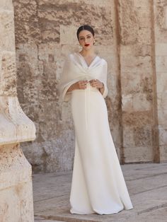 a woman standing in front of a stone wall wearing a white dress and shawl