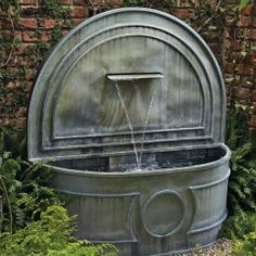 a water fountain in the middle of a garden with plants around it and a brick wall behind it