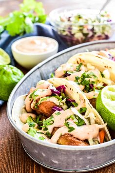 two tacos in a metal bowl on a wooden table with limes and other ingredients