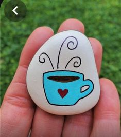 a hand holding a painted rock with a cup of coffee on it's side