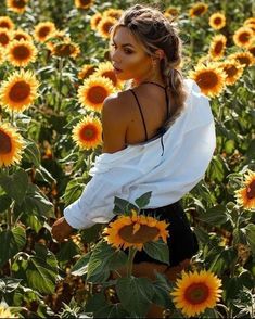 a woman standing in a field of sunflowers wearing a white blouse and black shorts