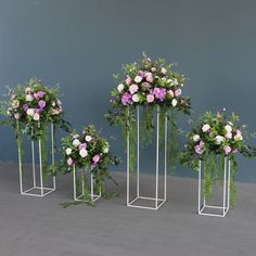 three metal plant stands with flowers and greenery in them on a concrete floor next to a blue wall