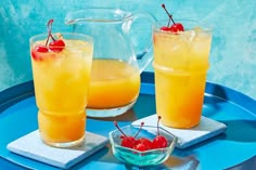 two glasses filled with orange juice and cherries on a tray next to a pitcher