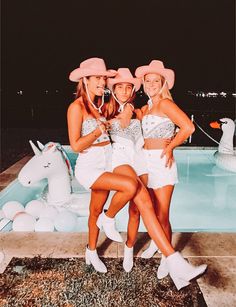 three women in white outfits and pink hats posing by a pool with an unicorn statue