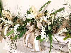 an arrangement of white flowers and greenery on a mantel with burlap ribbon