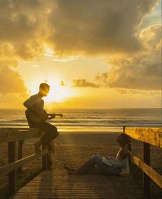 two people sitting on a wooden bench near the beach at sunset with an acoustic guitar