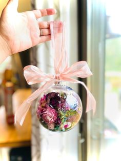 a hand is holding a glass ornament with flowers in it and pink ribbon