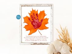 an orange maple leaf with hand prints on it in front of a white frame and dried grass